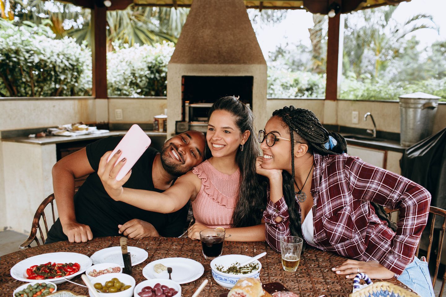 Three friends smiling together for a selfie while sharing food at an outdoor table